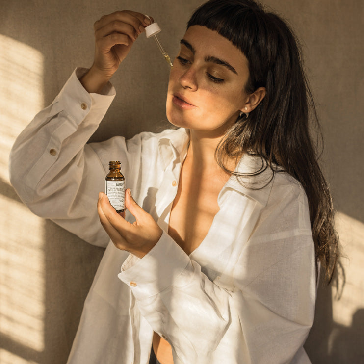 Woman holding a dropper above her cheek while holding a bottle labeled '004 DOPE your wrinkles FACE Oil SERUM, Olive Tree People'.
