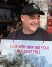 Hombre sonriente sosteniendo un cartel que dice “YOUR VERY OWN 100 YEAR OLD OLIVE TREE” frente a una cafetería