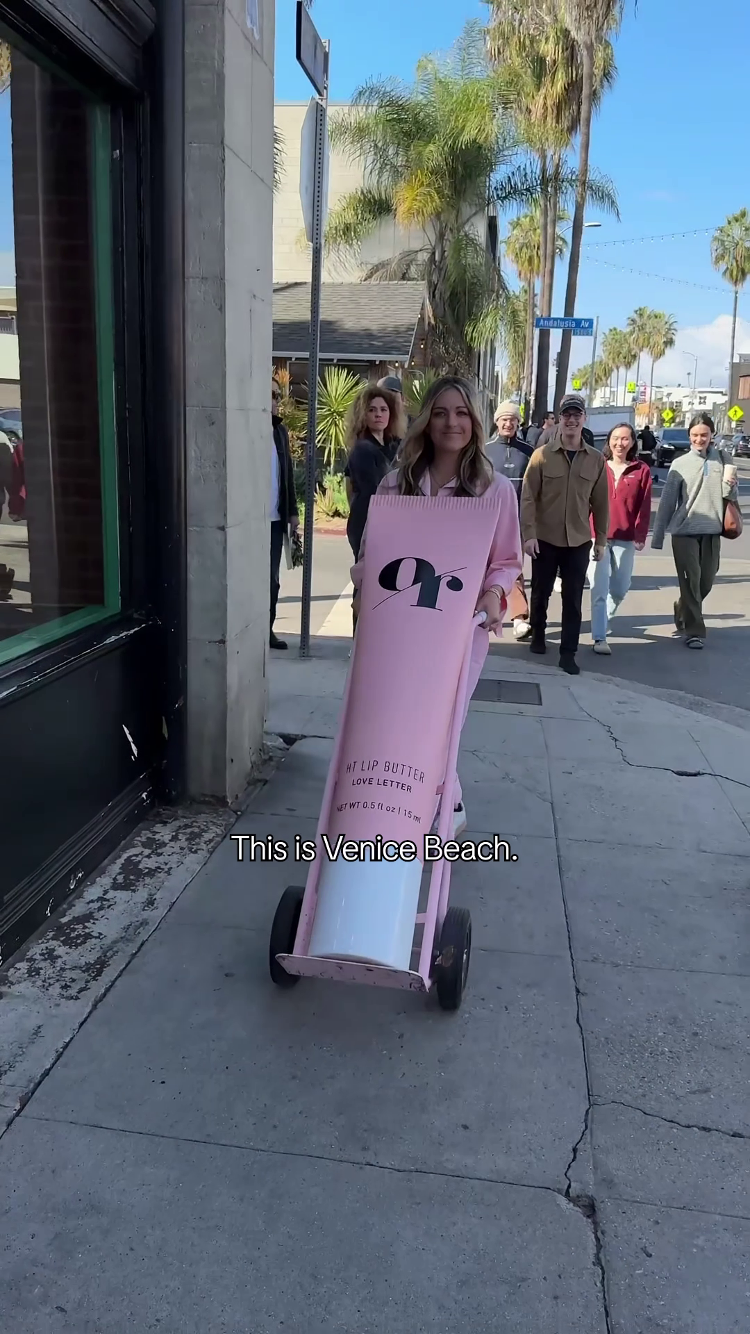 Woman pushing giant pink lip-balm prop on a dolly along a Venice Beach sidewalk, overlaid text: This is Venice Beach