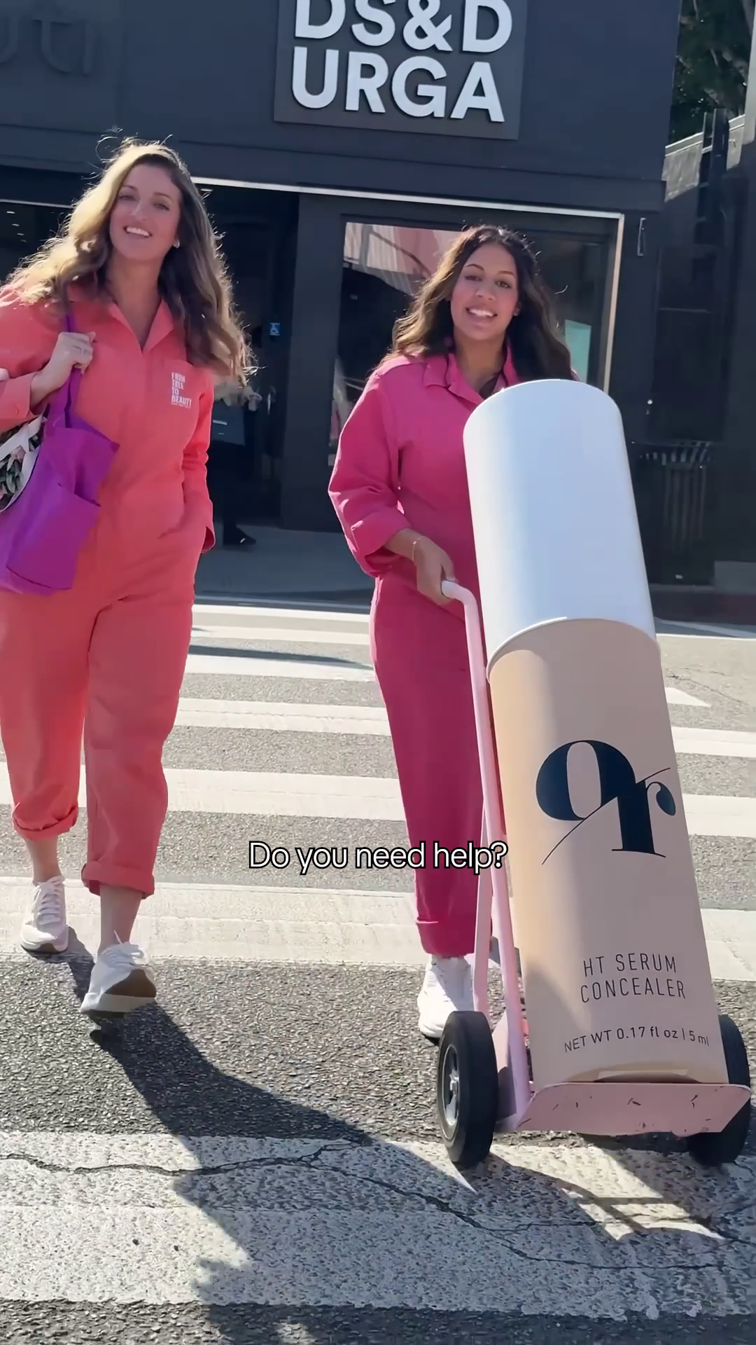 Two women pushing HT SERUM CONCEALER on cart at crosswalk; sign DS&D URGA; Do you need help? NET WT 0.17 fl oz 5 ml
