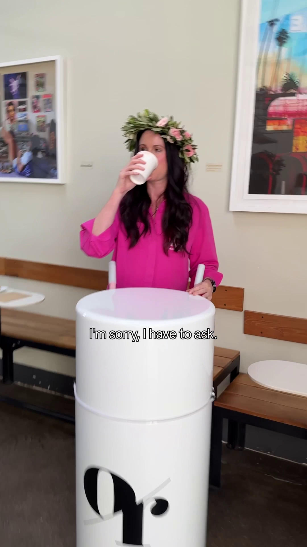 Woman in pink blouse with flower crown drinking from paper cup behind a white cylinder. Text reads I'm sorry, I have to ask.