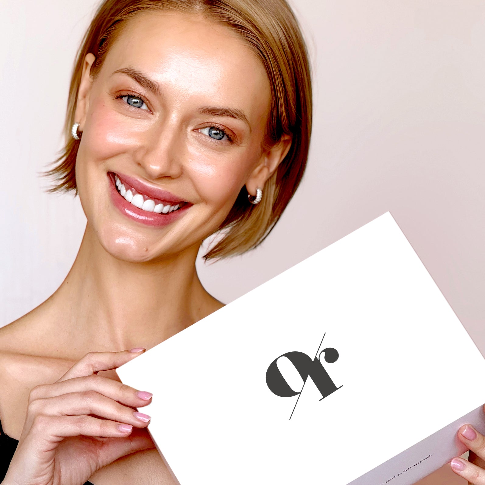 Smiling model holding a white Olive Tree People gift box with a black stylized logo against a pale pink background