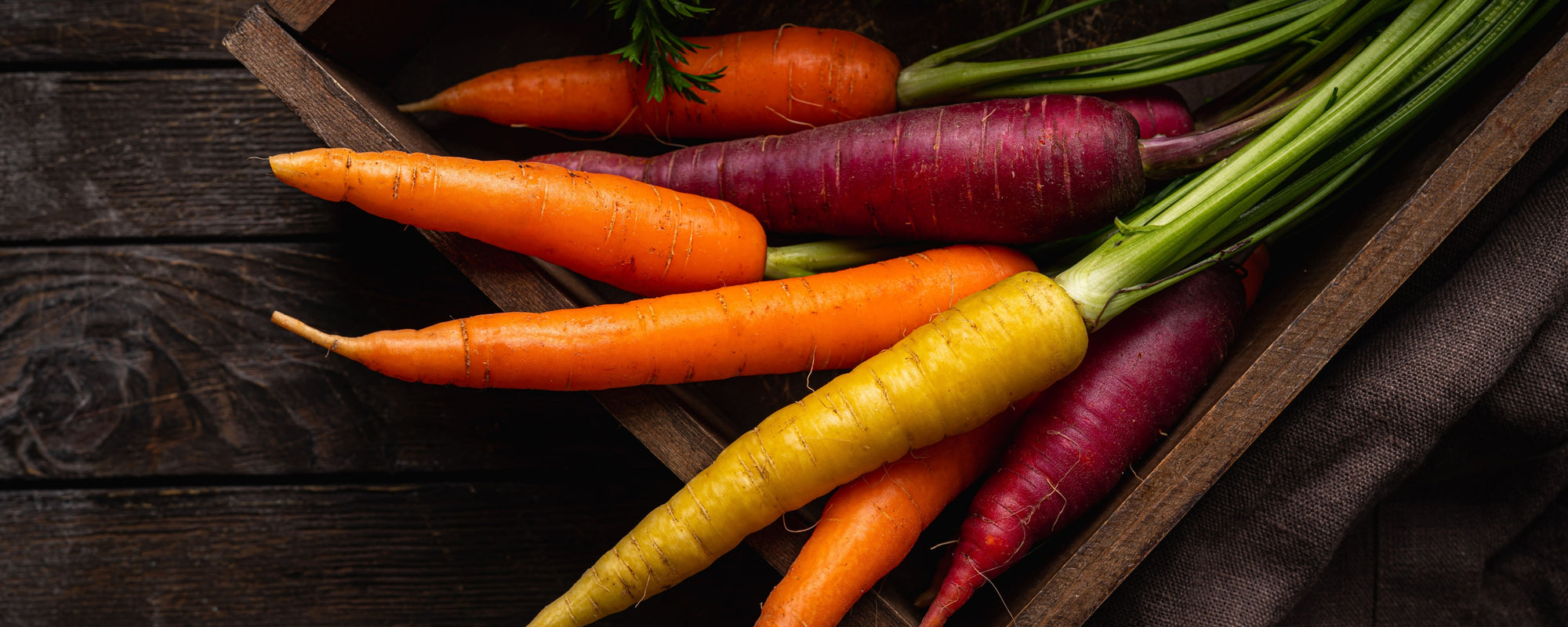 Multicolored carrots (orange, purple, yellow) with greens in a wooden crate on dark wood background