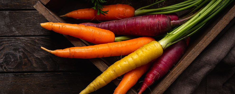 Multicolored carrots (orange, purple, yellow) with greens in a wooden crate on dark wood background