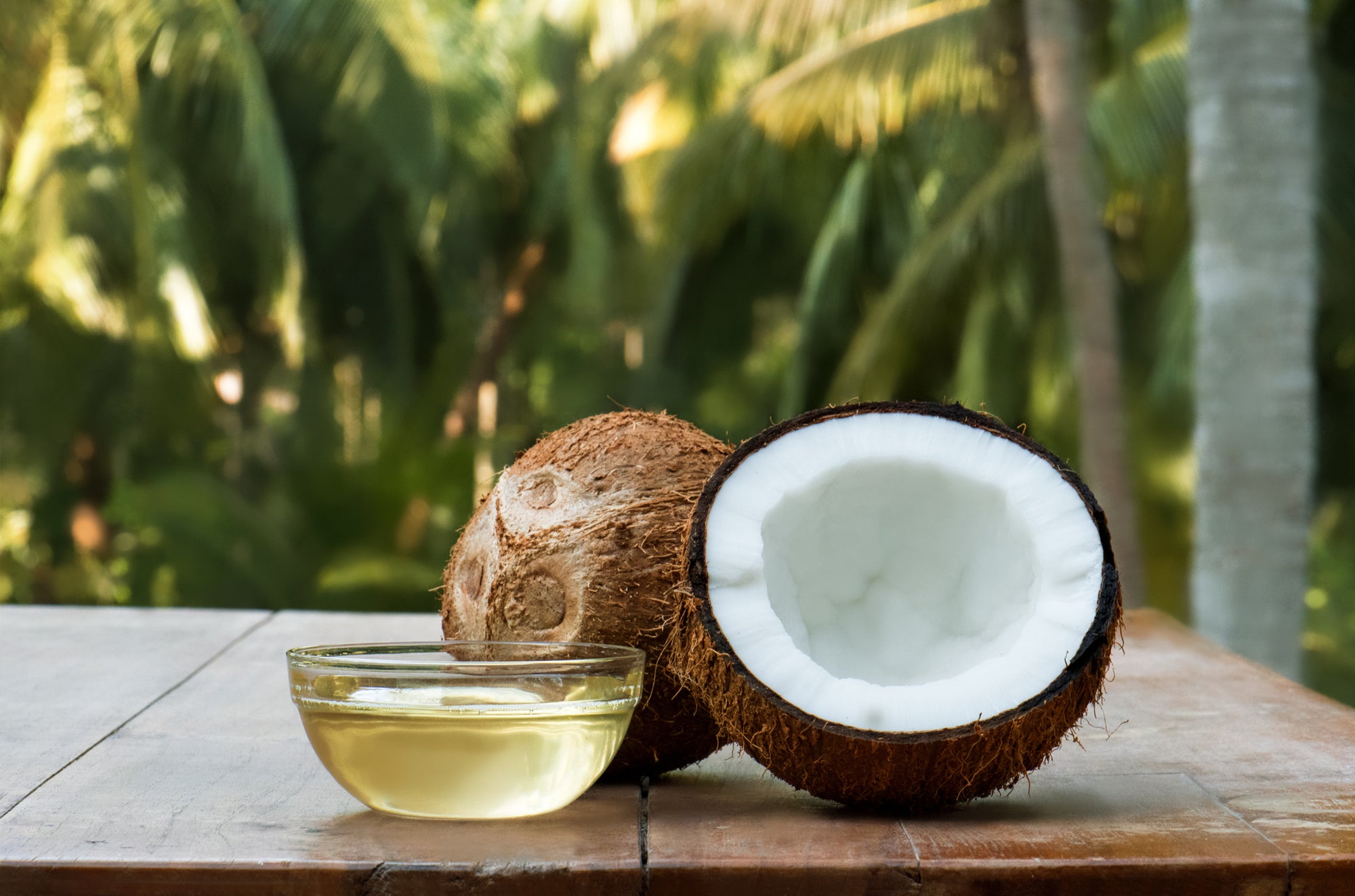 Halved coconut and whole coconut beside a glass bowl of coconut oil on a wooden table with palm trees in the background