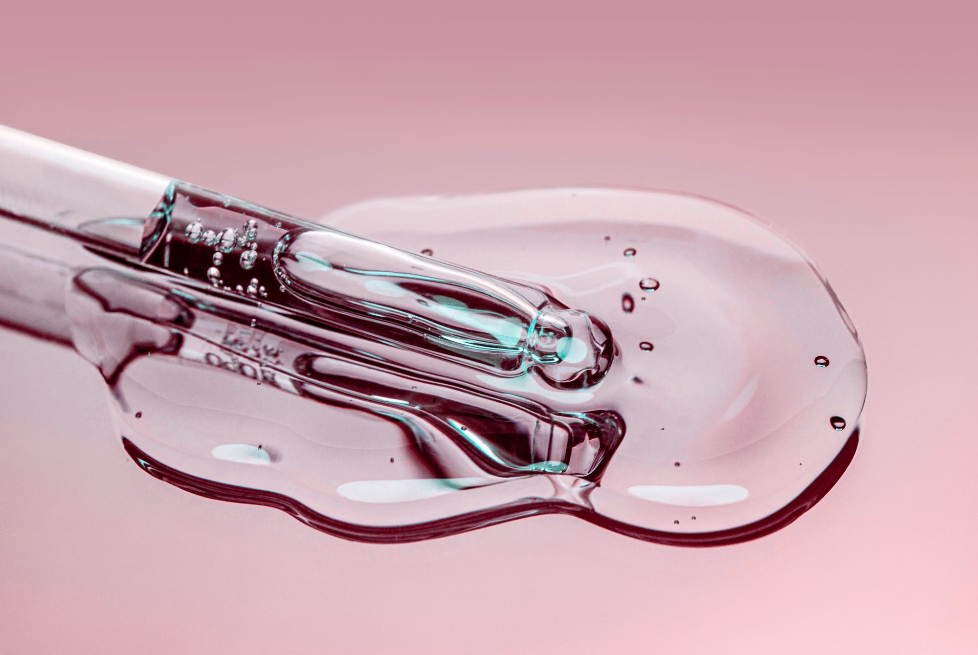 Glass dropper with clear serum pooled on a pink surface, close-up showing droplets