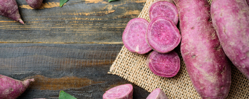 Purple sweet potatoes, whole and sliced, arranged on burlap atop a wooden table.