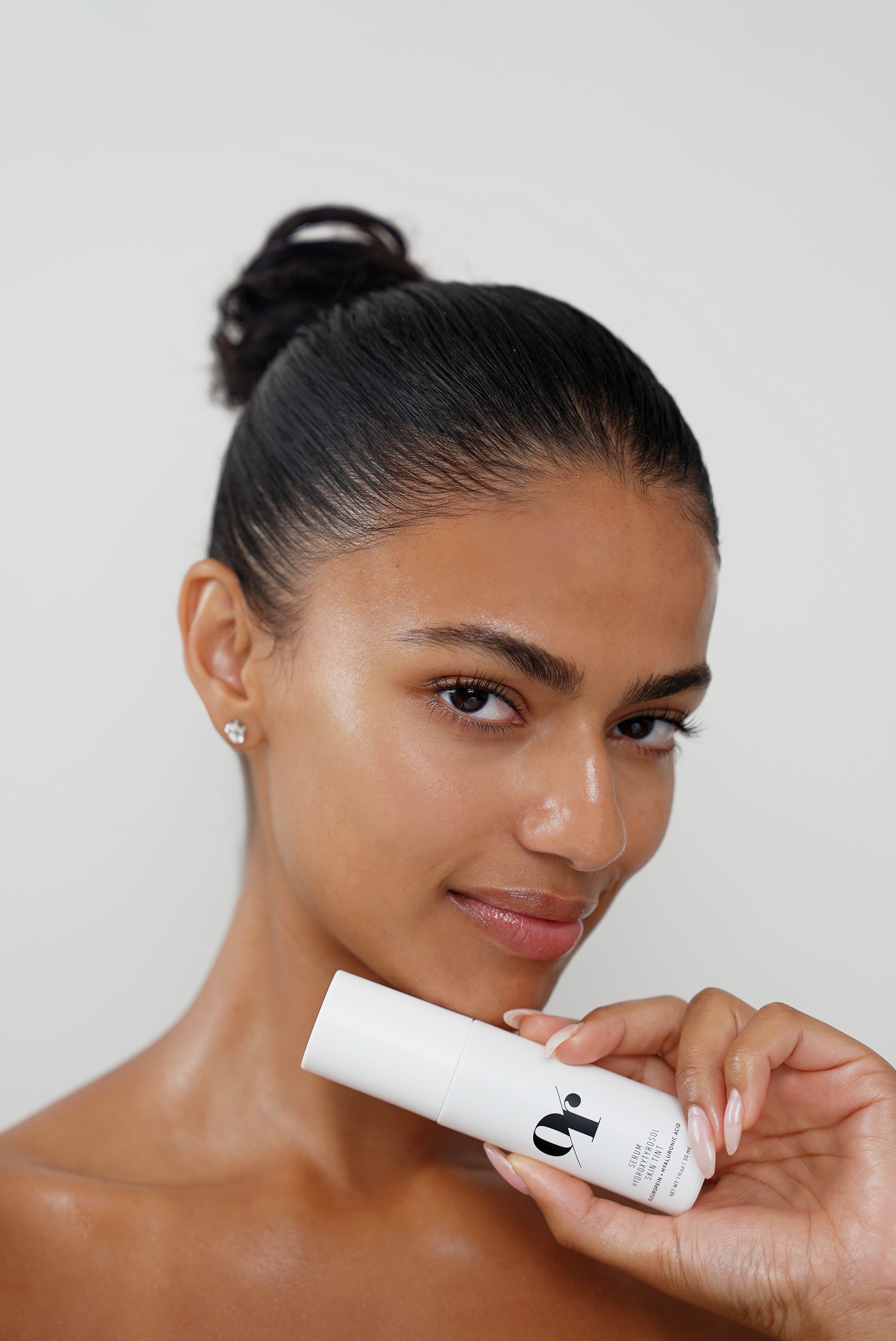 Woman with slicked-back bun holding white bottle labeled 'or' near her jaw; dewy skin and diamond stud earring.