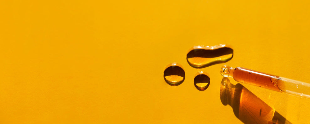 Glass dropper with amber liquid and three droplets on a bright yellow background.