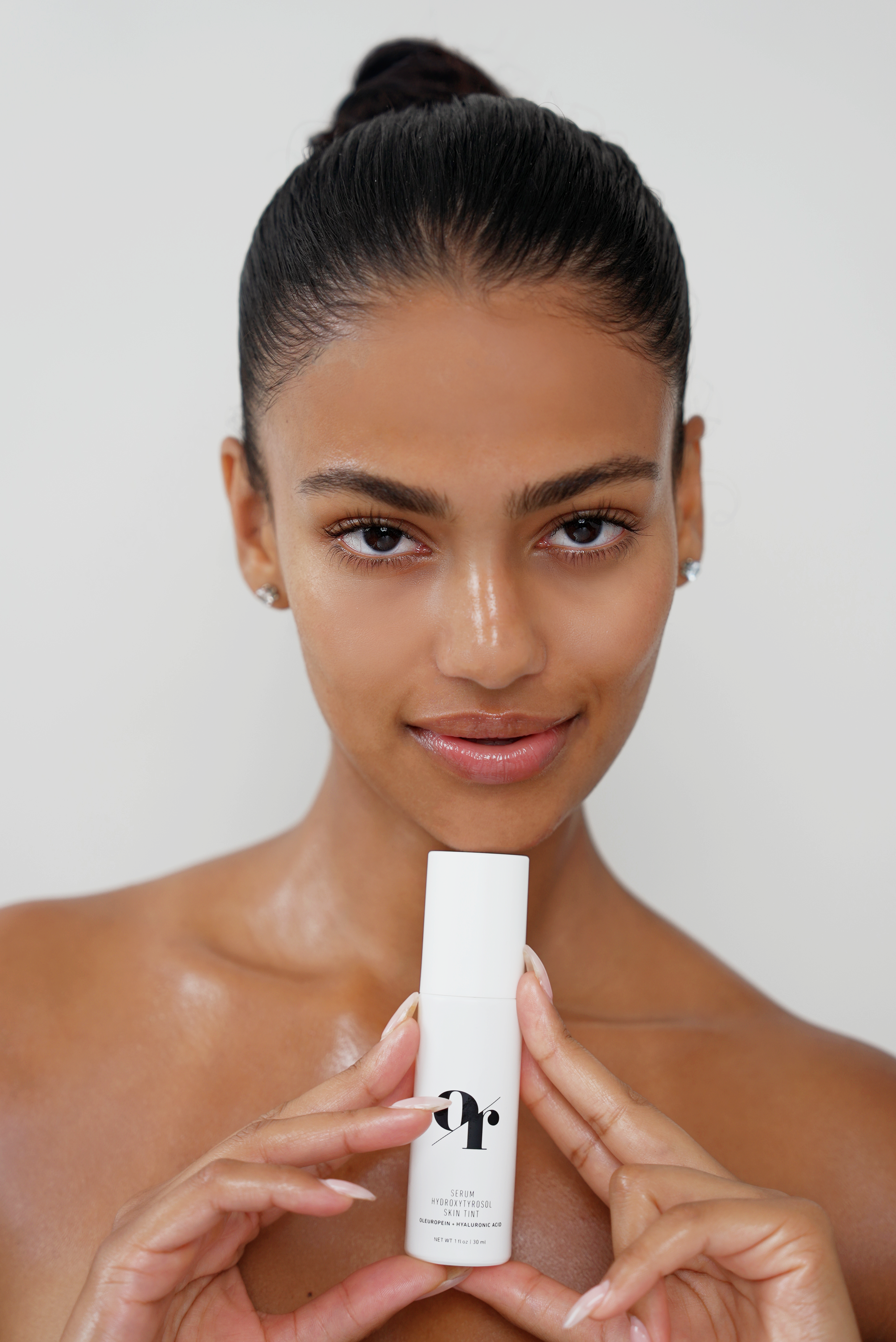 Woman holding white bottle labeled 'pr.' — SERUM HYDROXYTROSOL SKIN TINT OLEUROPEIN + HYALURONIC ACID NET WT 1 fl oz 30 ml