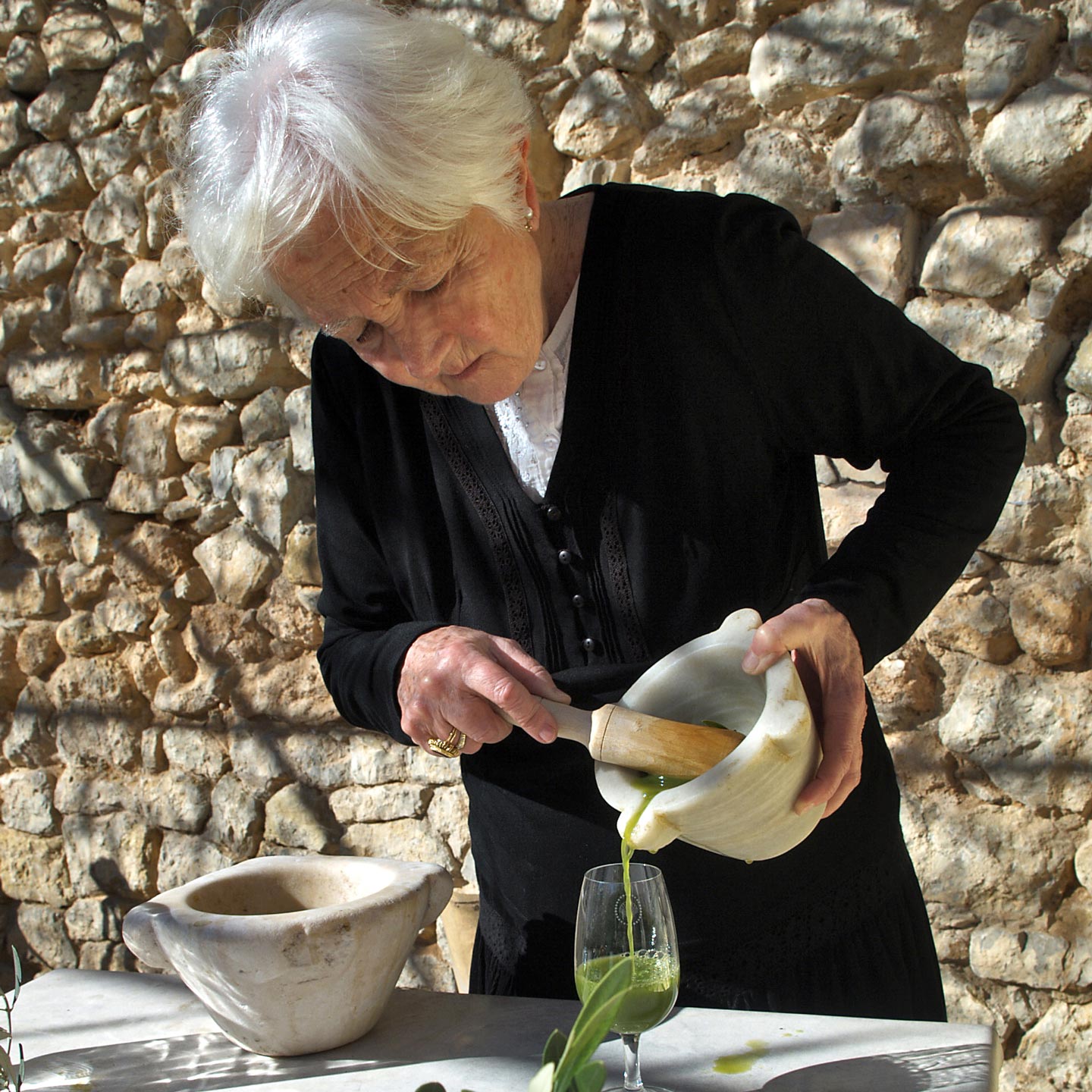 Elderly woman pouring freshly pressed green olive oil from a marble mortar into a glass, stone wall background