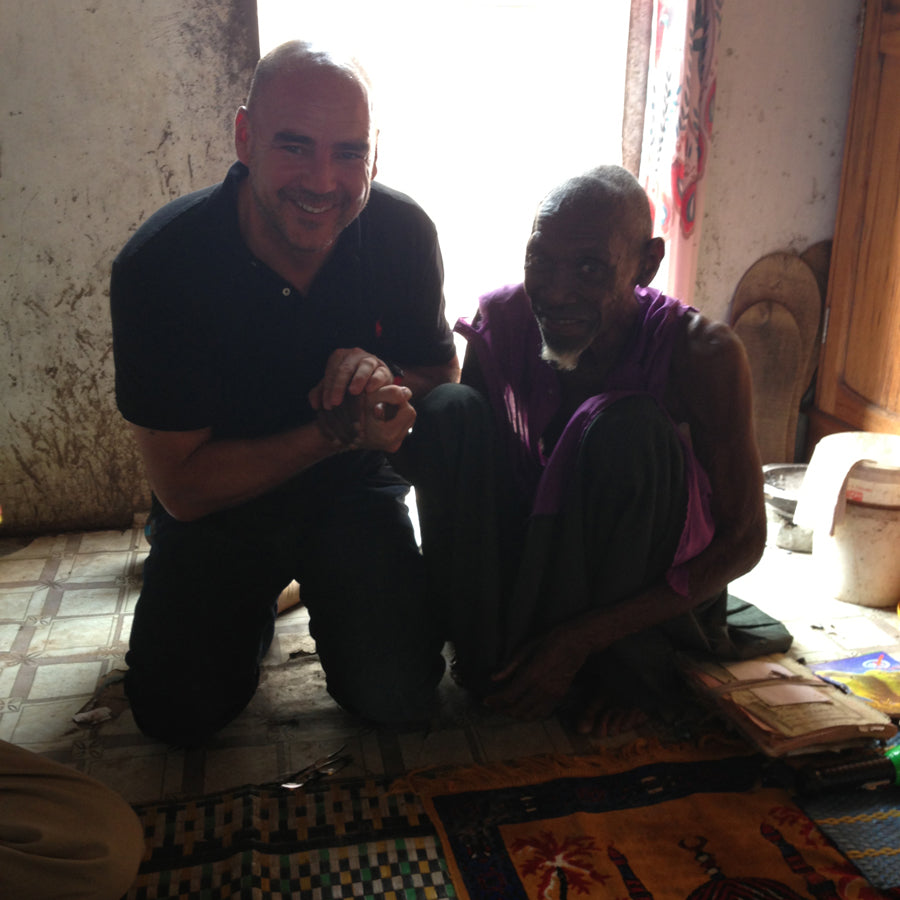 Smiling man kneeling and holding hands with an elderly man seated on a mat inside a modest, sunlit room.