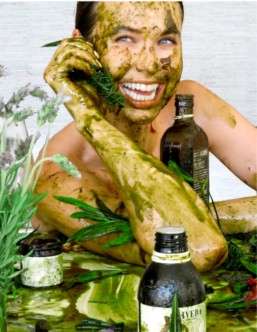 Woman smiling covered in green herbal facial and body mask, holding a rosemary sprig beside AVEDA bottles and jars on a reflective surface.