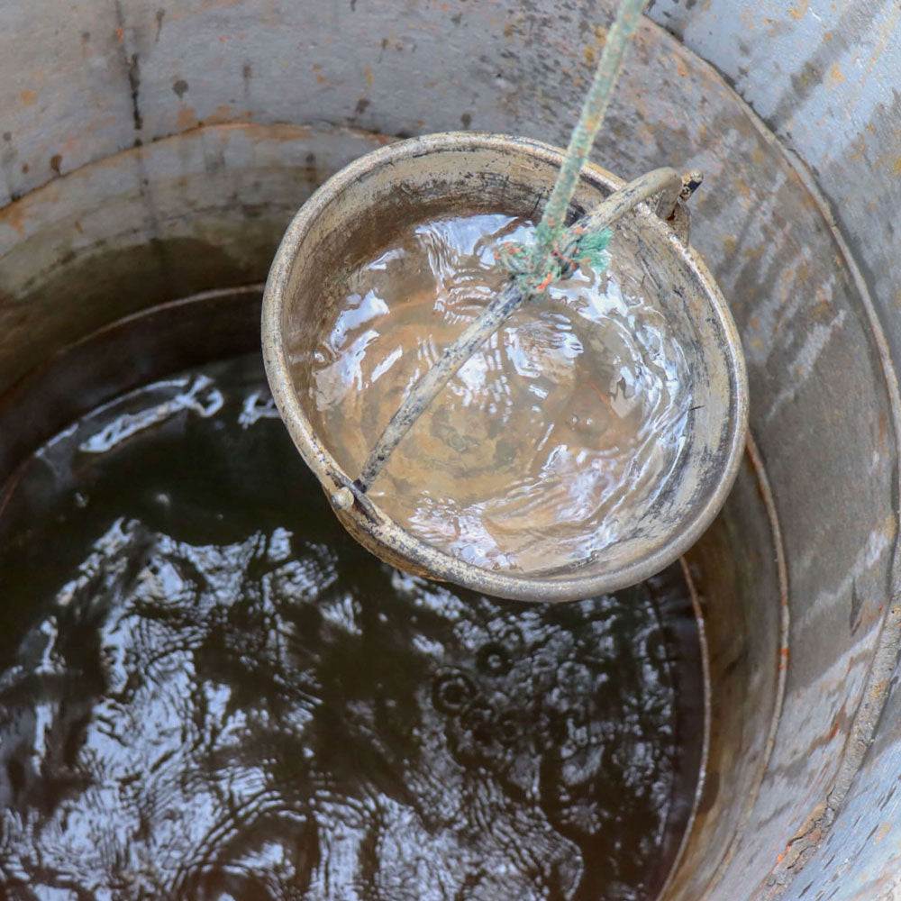 Rusty metal bucket suspended by a rope over a well, half-filled with water showing ripples and reflections.